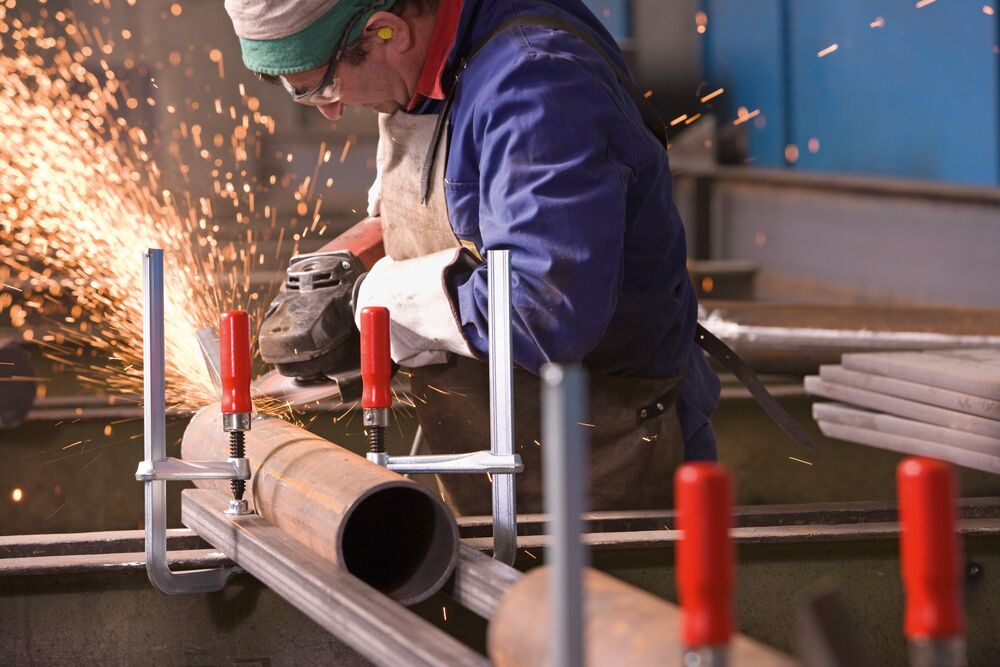 Des serre-joints entièrement en acier avec poignée en bois serrent une structure en acier, un individu travaille avec une meuleuse d'angle